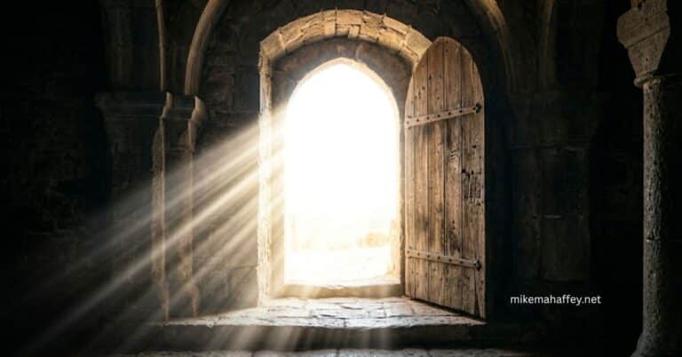 A heavy wooden door stands open in an ancient stone archway as brilliant rays of white-gold light pour into a dark stone interior, illustrating the deliverance from Satan's power into the kingdom of Jesus Christ.