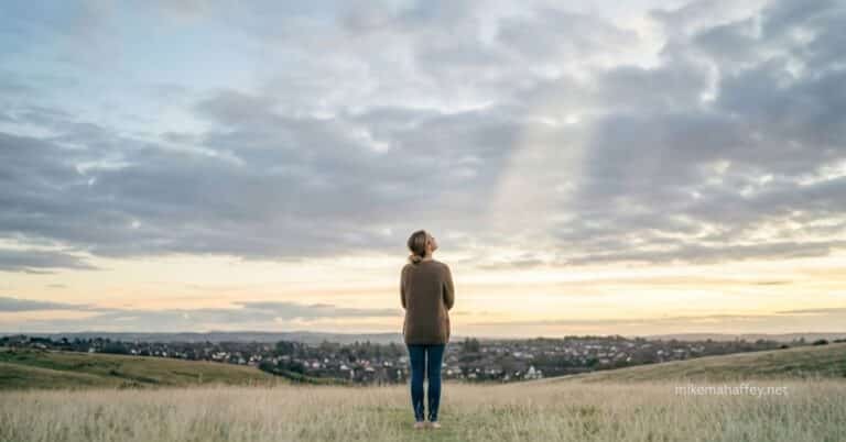 A woman stands alone on a grassy hillside looking up at a vast sky with a single ray of light breaking through the clouds, illustrating the question of whether God truly cares about personal struggles.