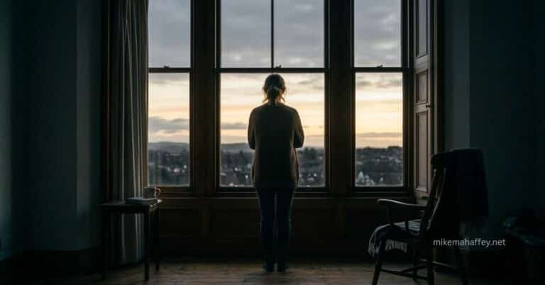 A woman stands alone in a dim room looking out a large window at a quiet dawn sky, reflecting the feeling of waiting on God in prayer.