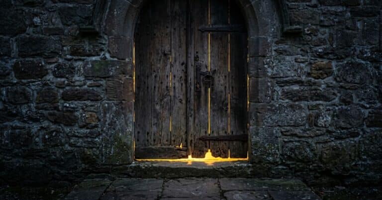 A weathered wooden door set in a dark stone wall with warm golden light glowing through the cracks and bottom edge, illustrating the truth of Jesus standing at the door and knocking in Revelation 3:20.