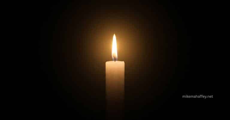 A single white candle burning steadily against a completely dark background, its warm golden flame radiating outward as a visual representation of the God is love meaning found in 1 John 4:8.