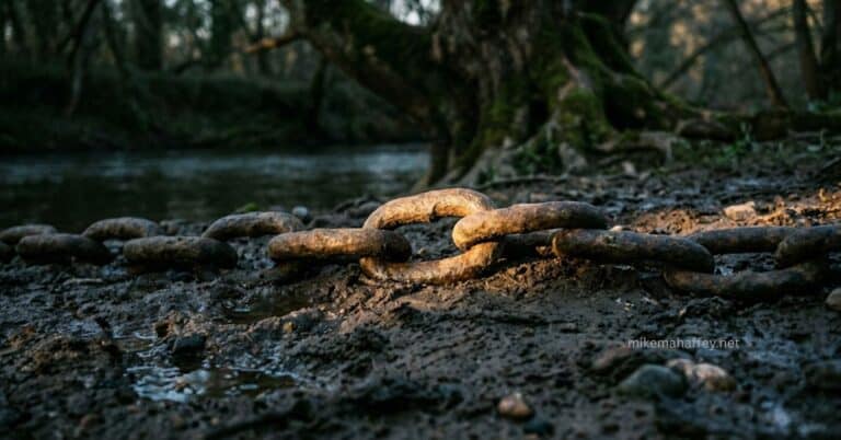 A photo of a heavy, rusted antique iron chain resting in the dark mud of a riverbank, with a warm light highlighting one main link, symbolizing the inherited sin problem mentioned in Romans 5:12.