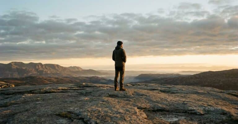 A solitary figure stands on solid stone at dawn with golden light breaking across the horizon, an image representing the settled peace with God that the blood of Jesus purchased at the Cross.