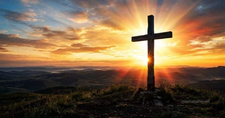 A wooden cross silhouetted against a crimson sunset sky with rays of light breaking through the clouds above, representing what the blood of Jesus accomplished at Calvary.
