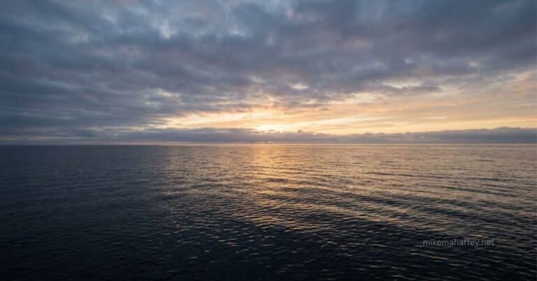 A calm dark sea reflects soft golden light breaking through clouds at the horizon, picturing the quiet, steady presence behind the Holy Spirit vs Spirit of God question.