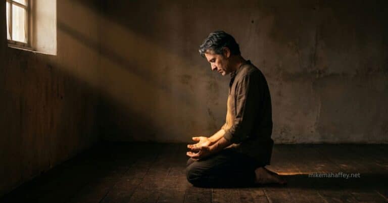A man kneeling on a wooden floor in a bare room, head bowed and hands open, with a shaft of light falling across him from a window — a picture of a believer standing in faith on the finished work of the blood of Jesus.