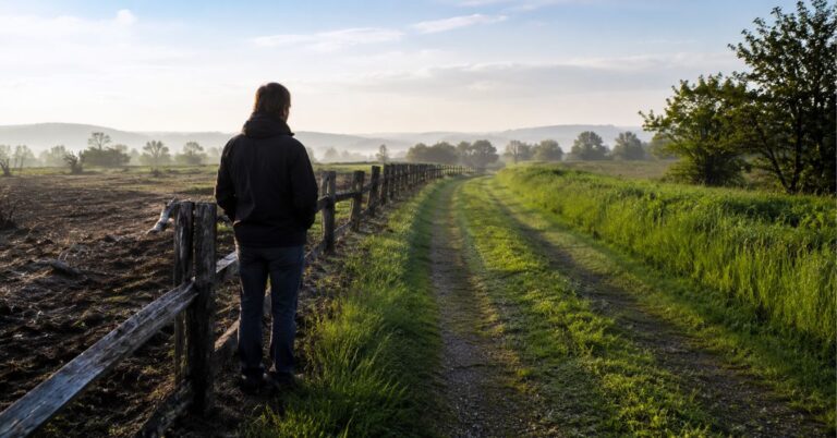Person standing between a barren field and a green countryside path in natural morning light.